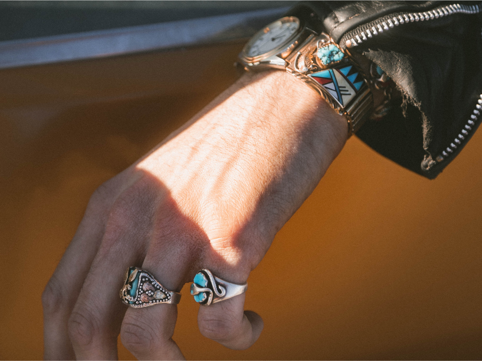 A hand hanging outside an orange car with two decorative rings and a watch.